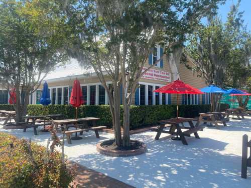 Outdoor dining area with picnic tables and colorful umbrellas near a restaurant, surrounded by trees and greenery.