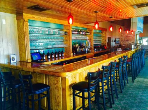 A wooden bar with red pendant lights, shelves of glasses, and various bottles lined up behind it. Empty bar stools are visible.