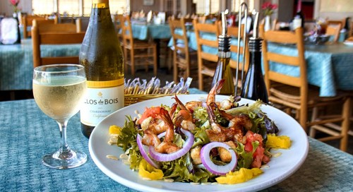 A plate of shrimp salad with vegetables, a glass of white wine, and a bottle on a table in a restaurant setting.
