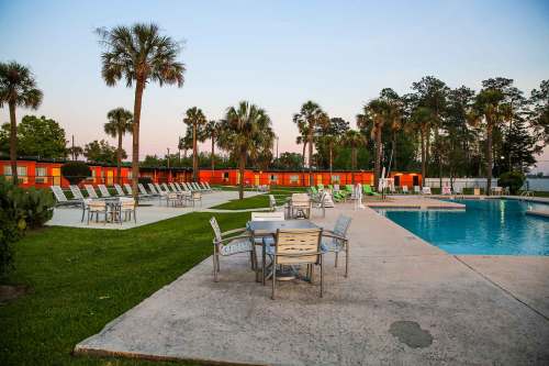 A serene pool area with lounge chairs, tables, and palm trees, surrounded by colorful buildings at sunset.