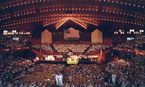A large choir performs in a grand hall with an American flag and illuminated signs, surrounded by an audience.
