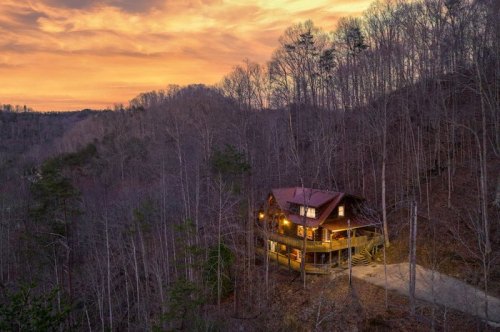 A cozy cabin nestled in a wooded area, illuminated at sunset with a colorful sky in the background.