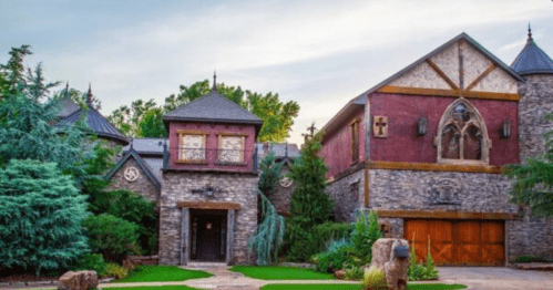 A large, ornate stone house with a mix of architectural styles, surrounded by lush greenery and a clear sky.