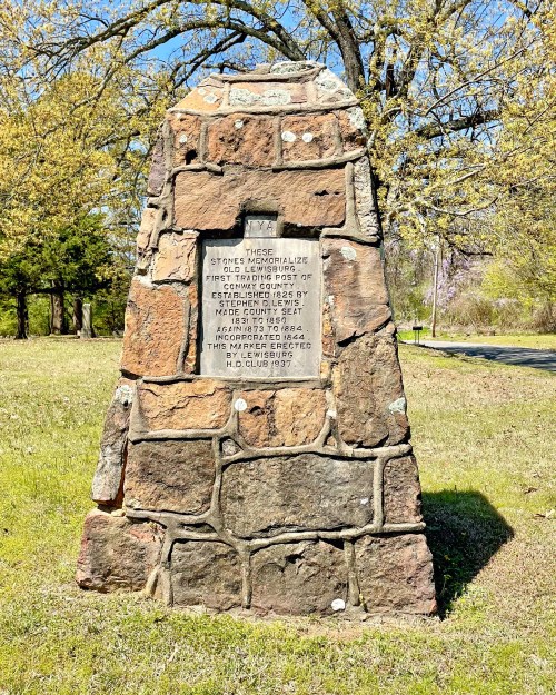 Stone monument with an inscription commemorating the first post office in the area, surrounded by grass and trees.