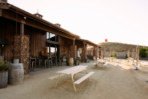 A rustic outdoor space with wooden tables, barrels, and a building featuring stone accents, set against a hillside.