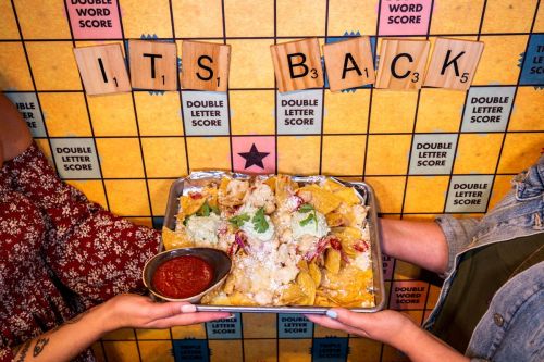 Two hands hold a tray of nachos topped with cheese and salsa, with a colorful game board backdrop spelling "IT'S BACK."