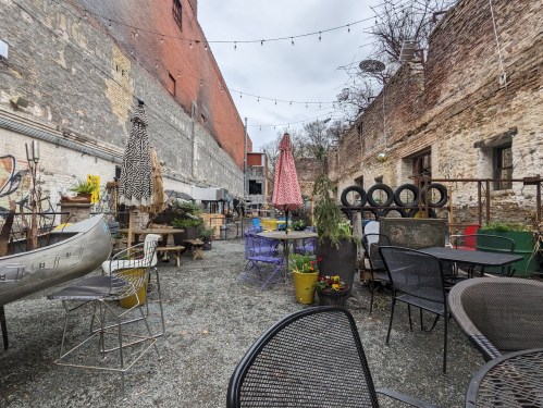 A rustic outdoor space with tables, chairs, umbrellas, and greenery, surrounded by brick walls and string lights.