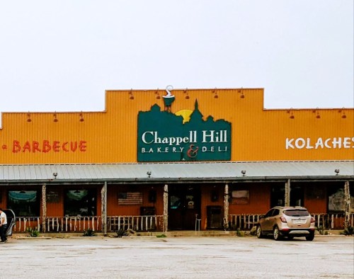 Exterior of Chappell Hill Bakery & Deli, featuring signs for barbecue and kolaches, with parked cars in front.