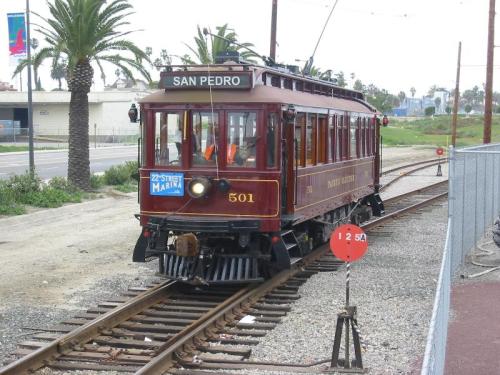 A vintage trolley labeled "San Pedro" on tracks, surrounded by palm trees and a cloudy sky.