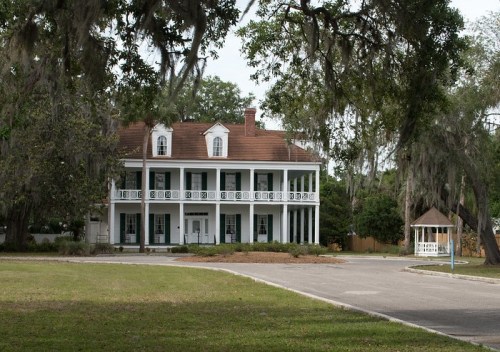A large, historic white house with a red roof, surrounded by trees and a grassy area, featuring a gazebo nearby.