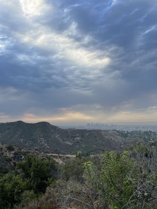 A scenic view of hills and the skyline of a city under a cloudy sky at dusk.
