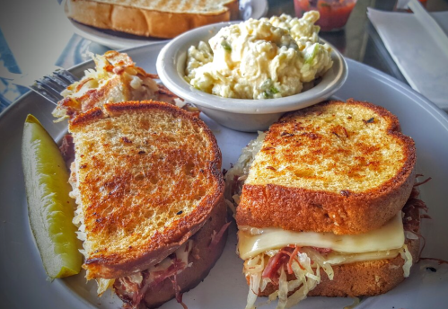 A plate with a grilled sandwich, pickles, and a side of potato salad. Toasted bread with melted cheese and sauerkraut.