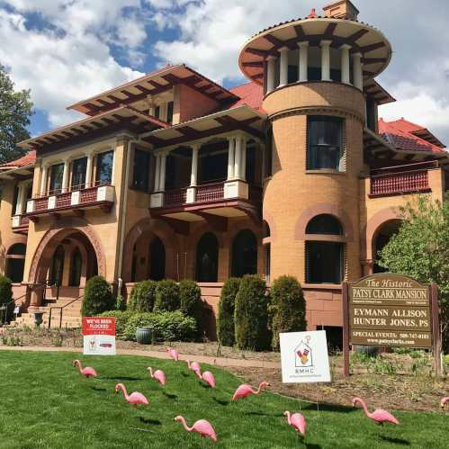 Historic Patsy Clark Mansion with pink flamingos in the foreground and event signs in a lush green yard.