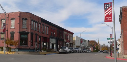 A quaint street in a small town featuring brick buildings, American flags, and parked cars under a blue sky.