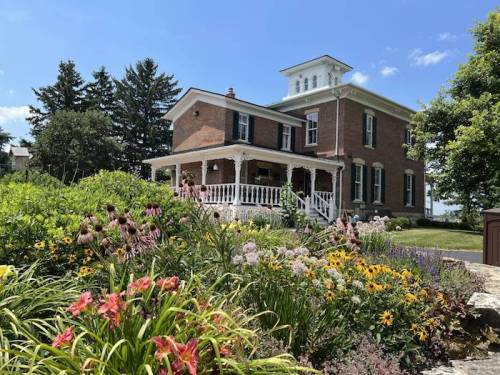 A charming brick house with a porch, surrounded by colorful flowers and greenery under a clear blue sky.