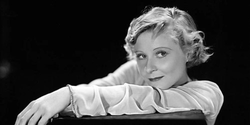 A vintage black-and-white portrait of a woman with short, wavy hair, resting her arms on a surface and smiling softly.