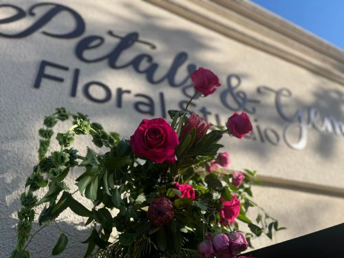 A close-up of vibrant pink flowers in front of a sign that reads "Petals & Gems Floral Studio."