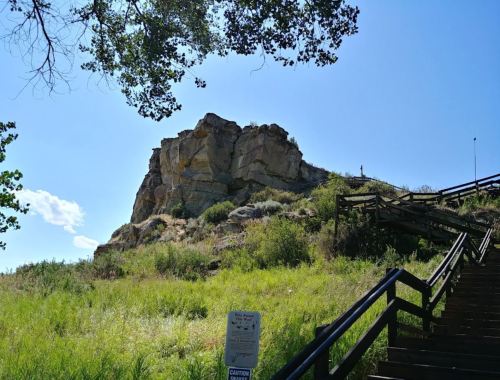 A rocky outcrop rises above a grassy area, with wooden steps leading up and trees framing the scene under a blue sky.