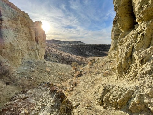 A sunlit canyon with rocky cliffs and a dry landscape under a blue sky, showcasing natural geological formations.