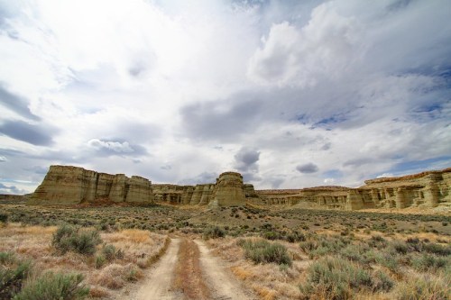 A dirt path leads through a vast landscape of cliffs and sagebrush under a cloudy sky.