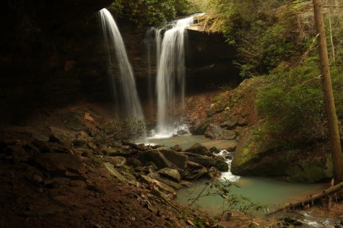 A serene scene of two waterfalls cascading into a tranquil pool, surrounded by lush greenery and rocky terrain.