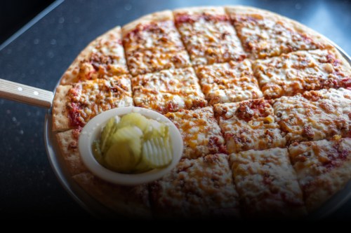 A freshly baked pizza with melted cheese, cut into squares, served with a side of pickles.