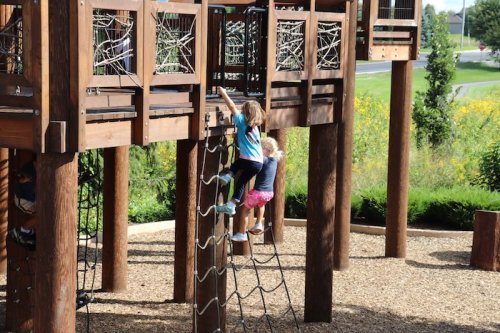 Two children play on a wooden playground structure, one climbing a rope net while the other watches nearby.