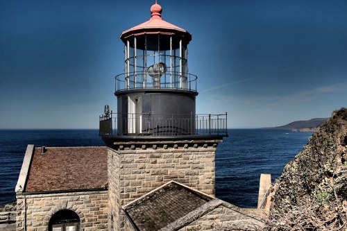 A historic lighthouse stands on a rocky coastline, overlooking the ocean under a clear blue sky.