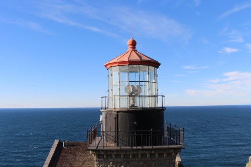 A lighthouse with a red roof stands against a clear blue sky and ocean backdrop.