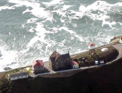Aerial view of a coastal property with houses and water towers near crashing waves along a rocky shoreline.
