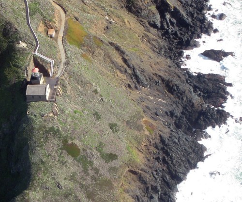 Aerial view of a lighthouse on a rocky coastline, surrounded by green vegetation and crashing waves below.