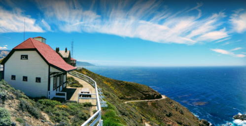 A coastal view featuring a white building with a red roof, overlooking the ocean under a blue sky with wispy clouds.