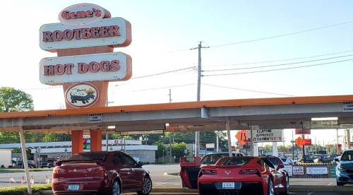 A retro sign for Gene's Rootbeer Hot Dogs, with cars parked underneath in a sunny setting.