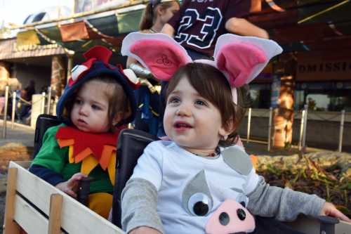 Two young children in costumes: one as a dragon and the other as a pig, sitting in a wagon at an outdoor event.