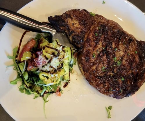 A grilled steak on a plate, accompanied by a fresh salad with greens, tomatoes, and a drizzle of dressing.