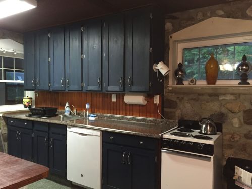 A cozy kitchen featuring dark blue cabinets, a stone wall, a sink, stove, and decorative items on the windowsill.
