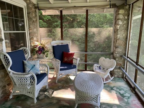 A cozy screened porch with wicker furniture, including chairs, a table, and a rocking chair, surrounded by greenery.