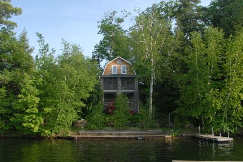 A lakeside house surrounded by lush green trees, with a wooden dock in the foreground and calm water.
