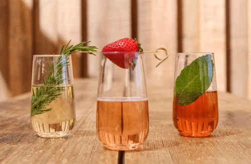 Three elegant glasses of colorful drinks, garnished with herbs and a strawberry, on a wooden table.