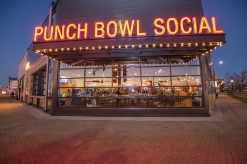 Exterior of Punch Bowl Social at dusk, featuring bright neon signage and large windows showcasing a lively interior.