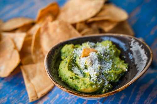 A bowl of guacamole topped with cheese, surrounded by crispy tortilla chips on a blue wooden surface.