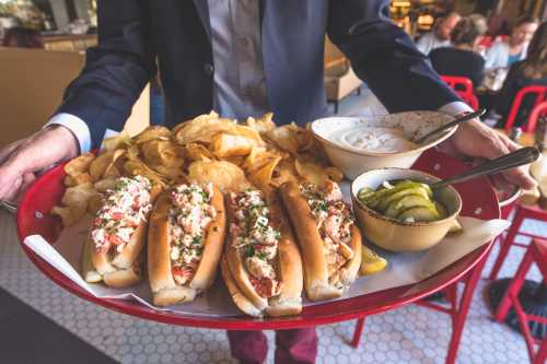 A waiter holds a tray with lobster rolls, potato chips, pickles, and dipping sauce in a restaurant setting.