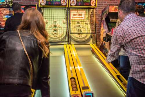 Two people playing skee-ball at an arcade, with colorful machines and a brick wall in the background.