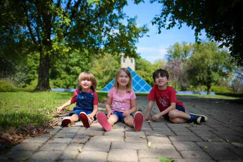 Three children sit on a paved path in a park, smiling and enjoying a sunny day with trees in the background.