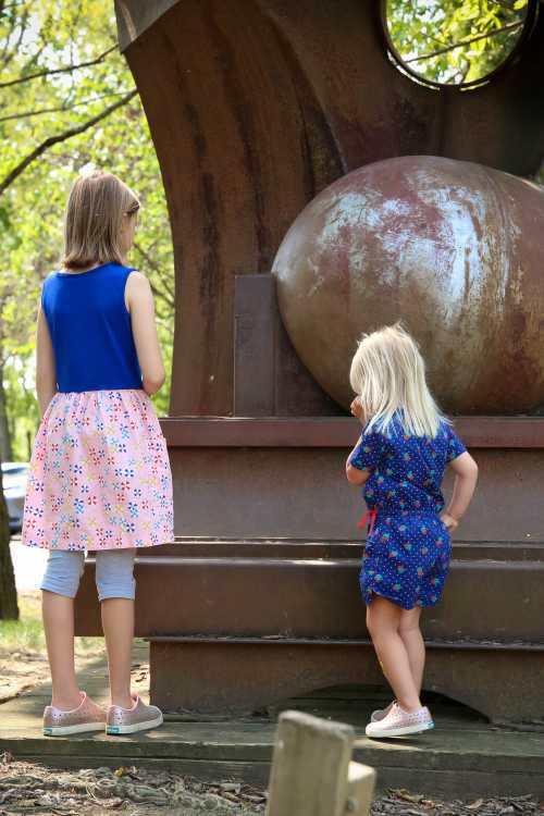 Two girls stand in front of a large metal sculpture, one wearing a blue top and floral skirt, the other in a blue dress.