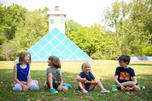 Four children sit on grass, smiling and chatting, with a glass pyramid structure and trees in the background.