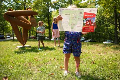 A child holds a map in front of a sculpture, with two other children playing in a grassy area surrounded by trees.