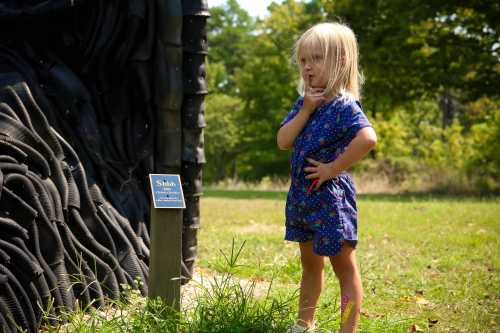 A young girl in a blue floral outfit stands thoughtfully next to a sign in a grassy area, with a textured black sculpture nearby.