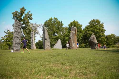 Children play among large stone sculptures in a grassy area, surrounded by trees and a clear blue sky.