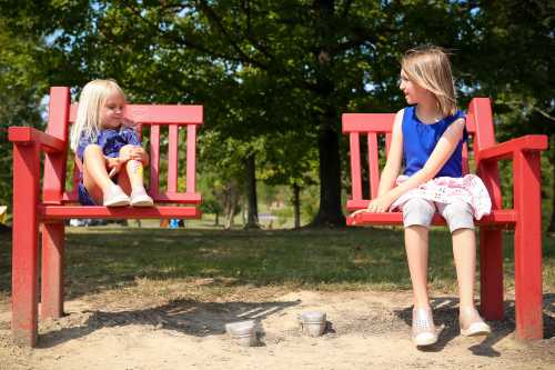 Two girls sit on red benches in a park, one smiling while the other looks at her, surrounded by trees and sand.